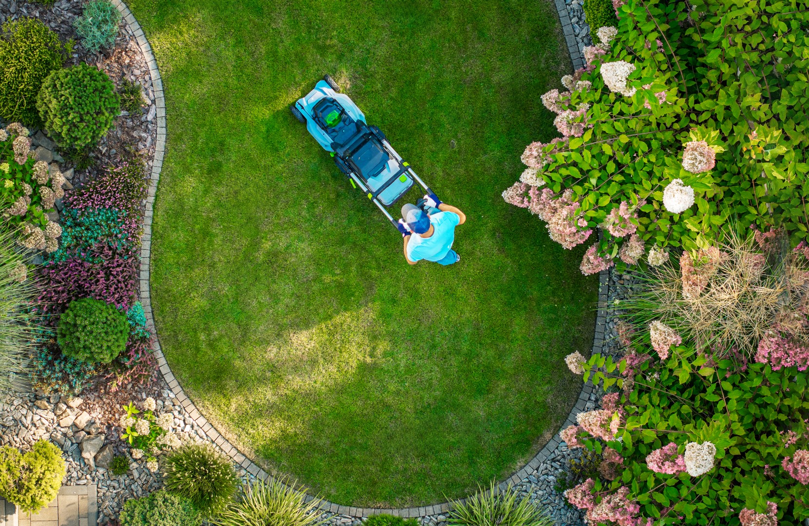 A person is mowing a perfectly shaped circular lawn in a vibrant garden filled with lush green plants and blooming flowers on a sunny day.